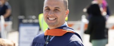 A man in a navy blue graduation gown and an orange sash smiles widely for the camera